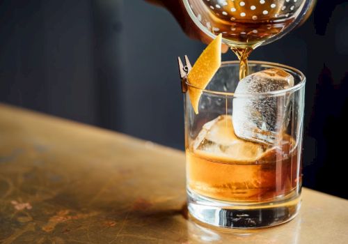 A whiskey glass with large ice cubes, an orange peel garnish, and a bubbly pour being topped from a decanter, on a wooden bar.