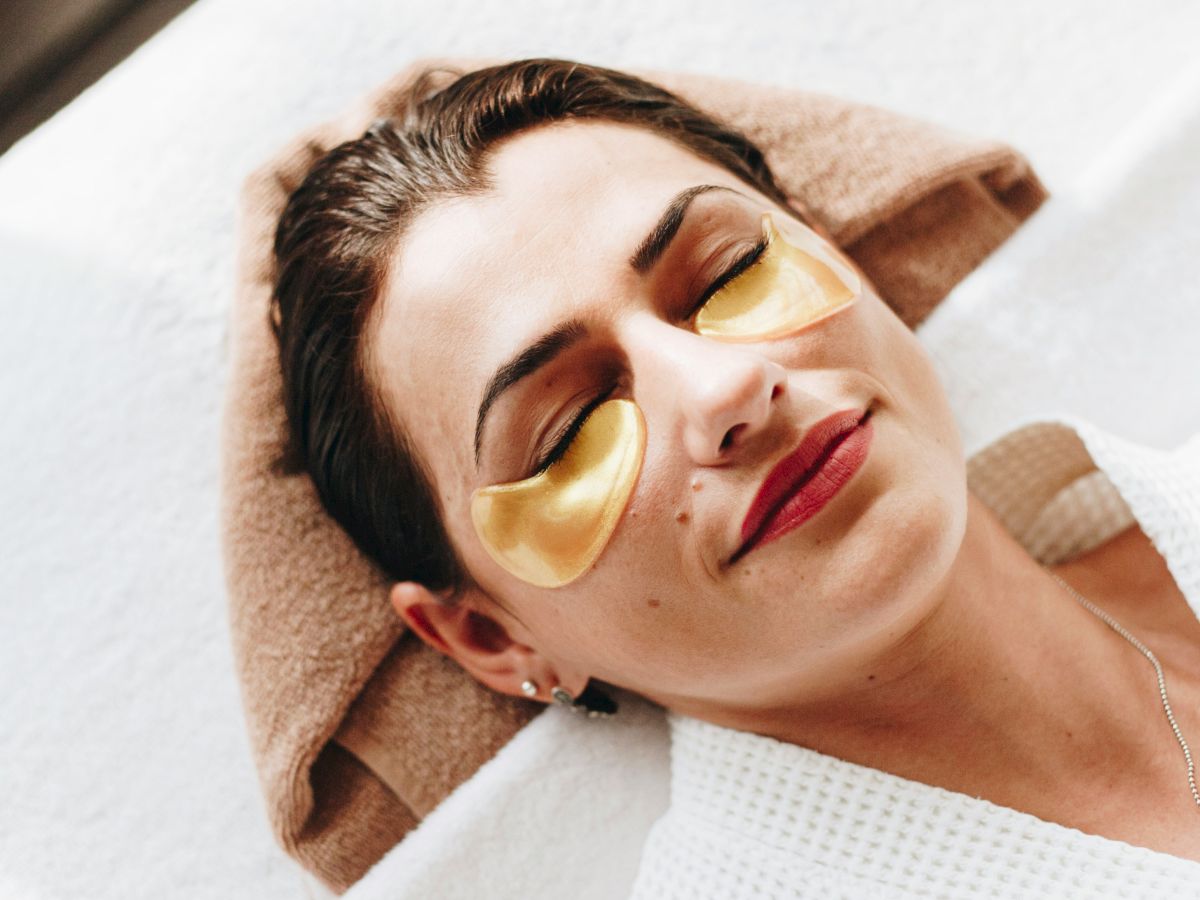 A person relaxing with golden under-eye patches, lying on a massage table with a towel under their head.
