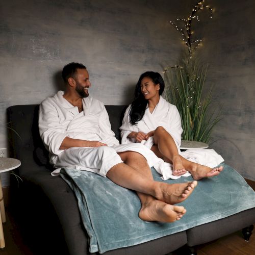 Two people lounge on a sofa in white robes, chatting warmly, with dim ambient lighting and plants around them.