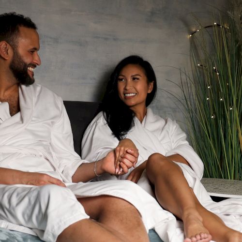 Two people in white robes sitting on a couch, smiling and holding hands in a spa-like setting.