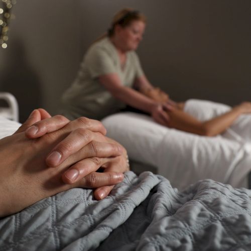 Two people hold hands in a cozy, dimly lit room, with someone receiving a massage in the background, set beside soft blankets and lights.