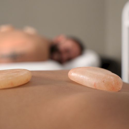 Two smooth stones resting on a bare back, blurred person lying on a bed in the background.