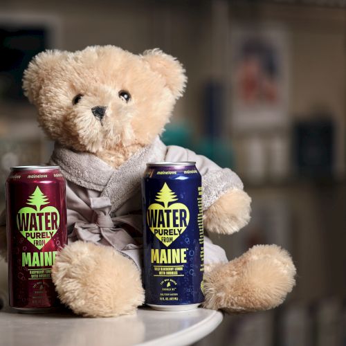 Two stuffed bears sit on a table with two cans of Maine Water Purity drink beside them.