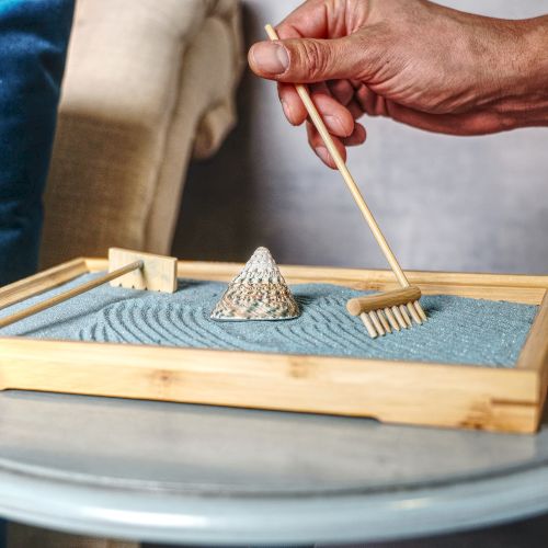 A wooden sand tray with a small cone, a tiny rake, and a raked pattern; a hand uses a stick to move the sand.