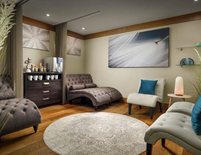 A cozy spa-like lounge with tufted chaise lounges, a rug, plants, soft seating, and calming wall art in a warm, modern setting.