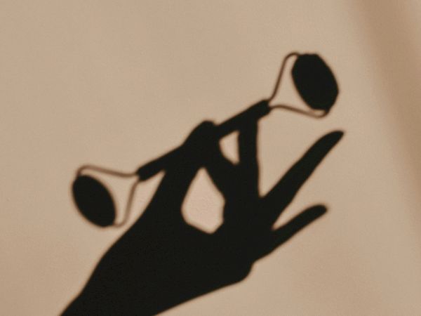 A black silhouetted hand balances two small glasses or cups on its fingers, against a light background, like a delicate juggling pose.