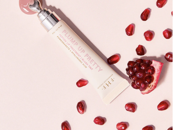 A PLUMPPURIFY PR&hellip; tube on a light background, scattered with red pomegranate seeds for a skincare/beauty ad.