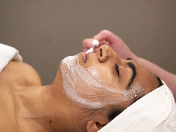 A person lying down with a facial mask being applied by a therapist, eyes closed, relaxed spa setting.