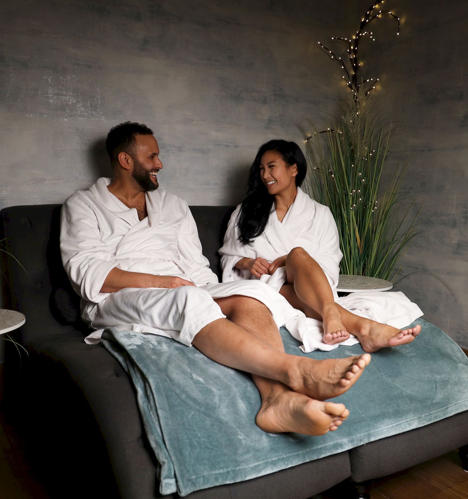 Two people sit on a sofa in robes, chatting softly in a cozy, dimly lit room with plants and a blanket, enjoying a relaxed spa vibe.