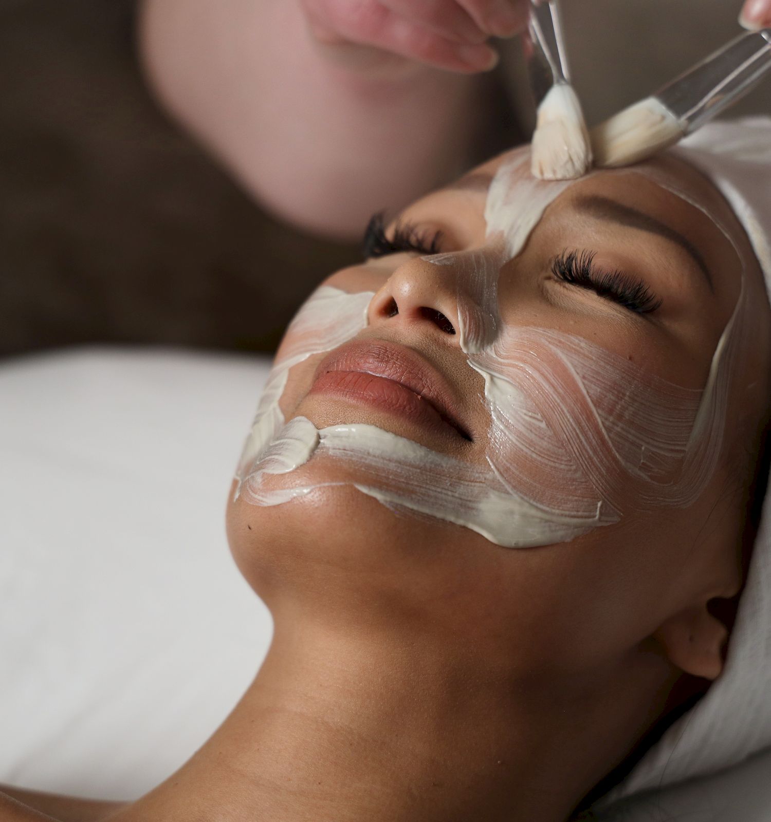 A woman lies with a white facial mask being applied, spa treatment in progress, relaxing beauty care.